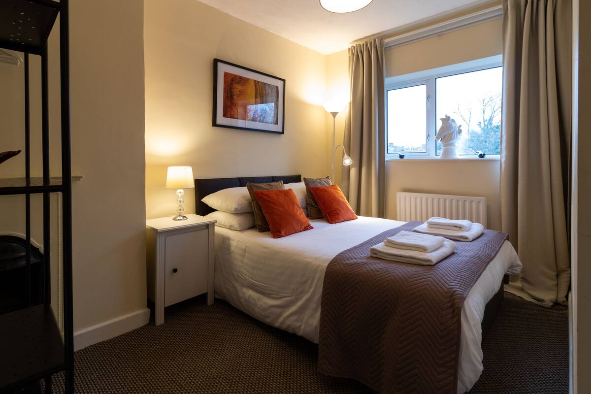 A hotel room with a bed, nightstand, lamp, window with curtains, and a radiator.