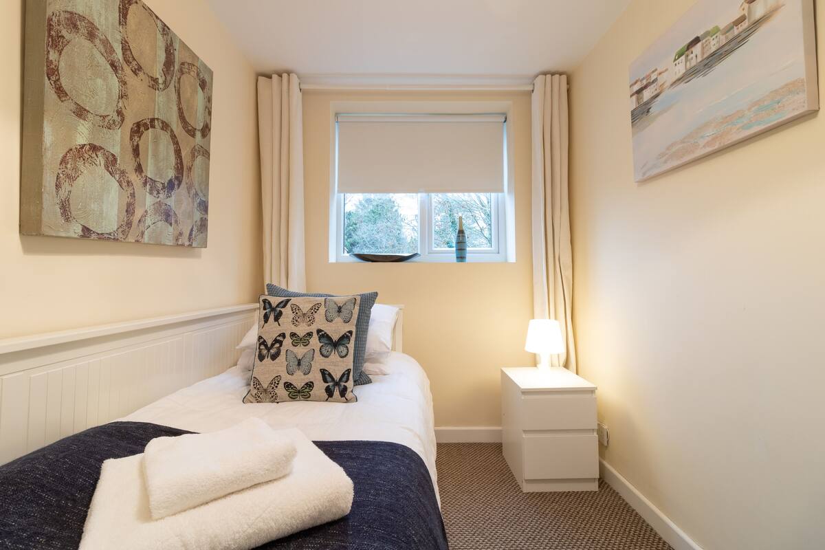 A neatly arranged bedroom with a bed, a nightstand, a window with curtains, and a painting on the wall.