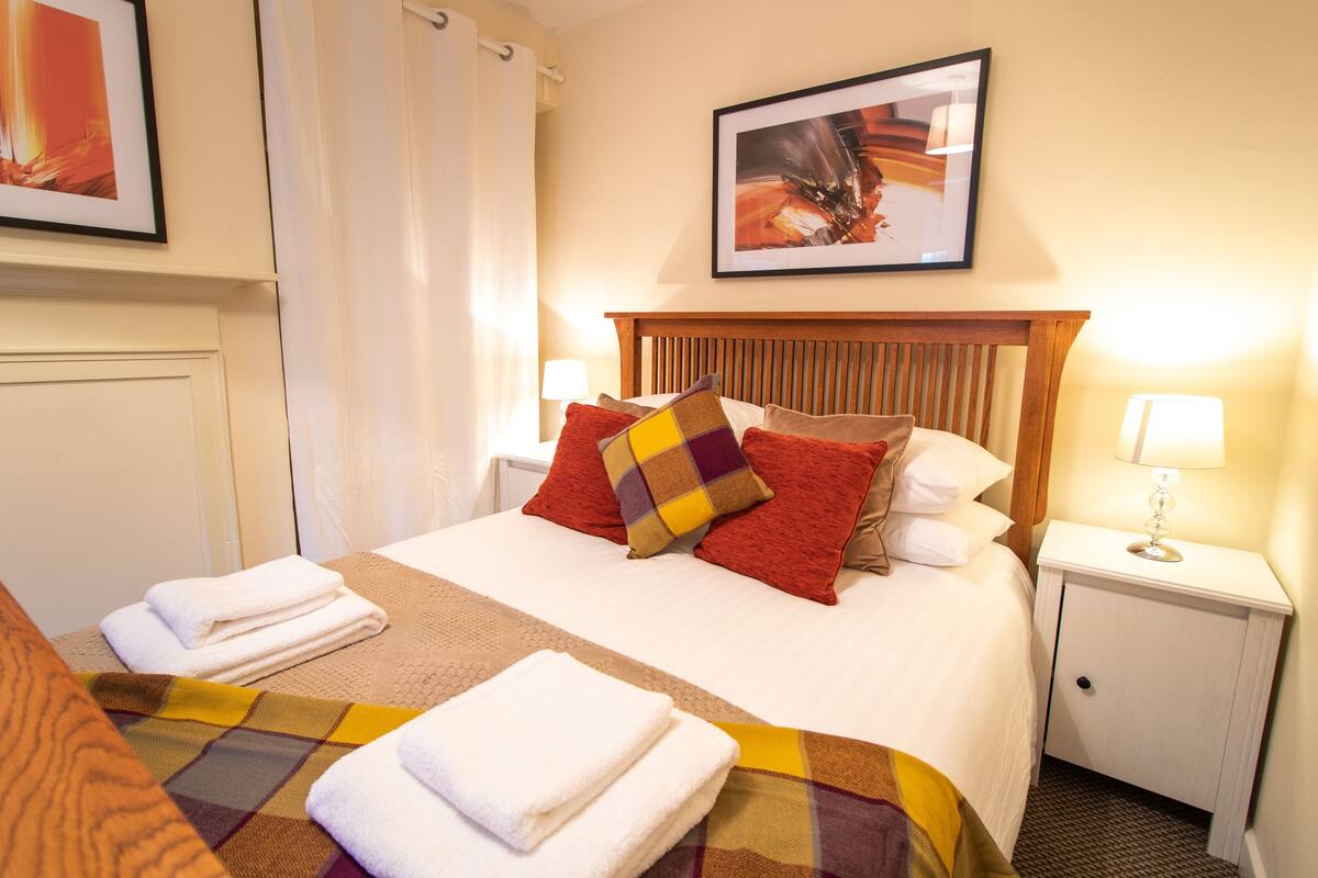 A neatly made bed with a plaid bedspread and multiple pillows, a small bedside table with a lamp, and a framed picture on the wall.