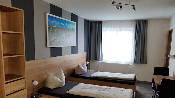 Une chambre d’hôtel avec deux lits simples, une tête de lit en bois, un tableau au mur, une fenêtre avec des rideaux et un bureau avec une chaise.