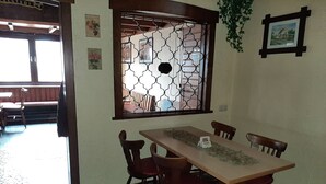 Un coin repas avec une longue table et quatre chaises, une fenêtre décorative ornée d’un motif géométrique et un tableau encadré accroché au mur.