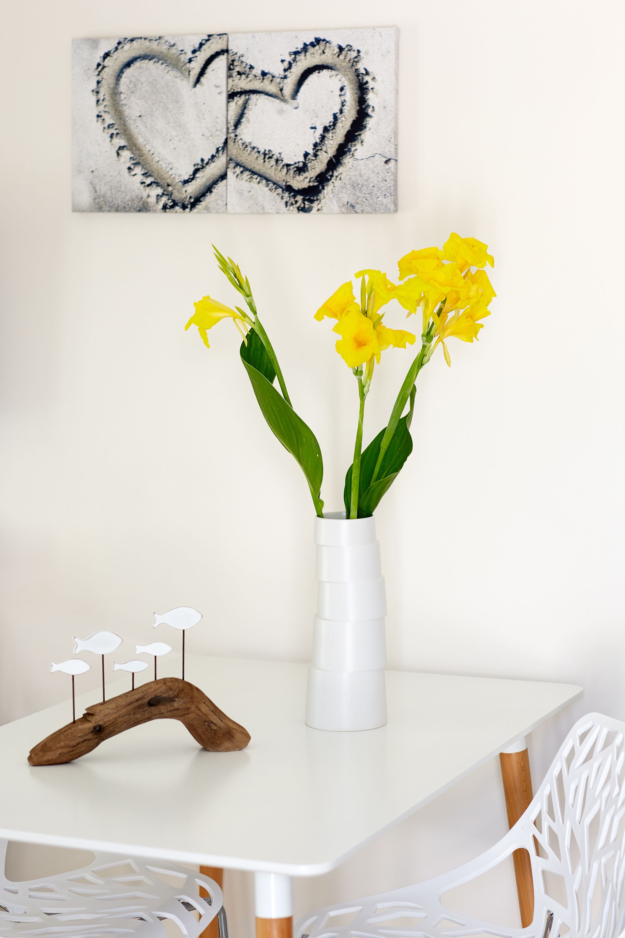 Una mesa blanca con un jarrón blanco que contiene flores amarillas, un objeto de madera sobre la mesa y una silla blanca.