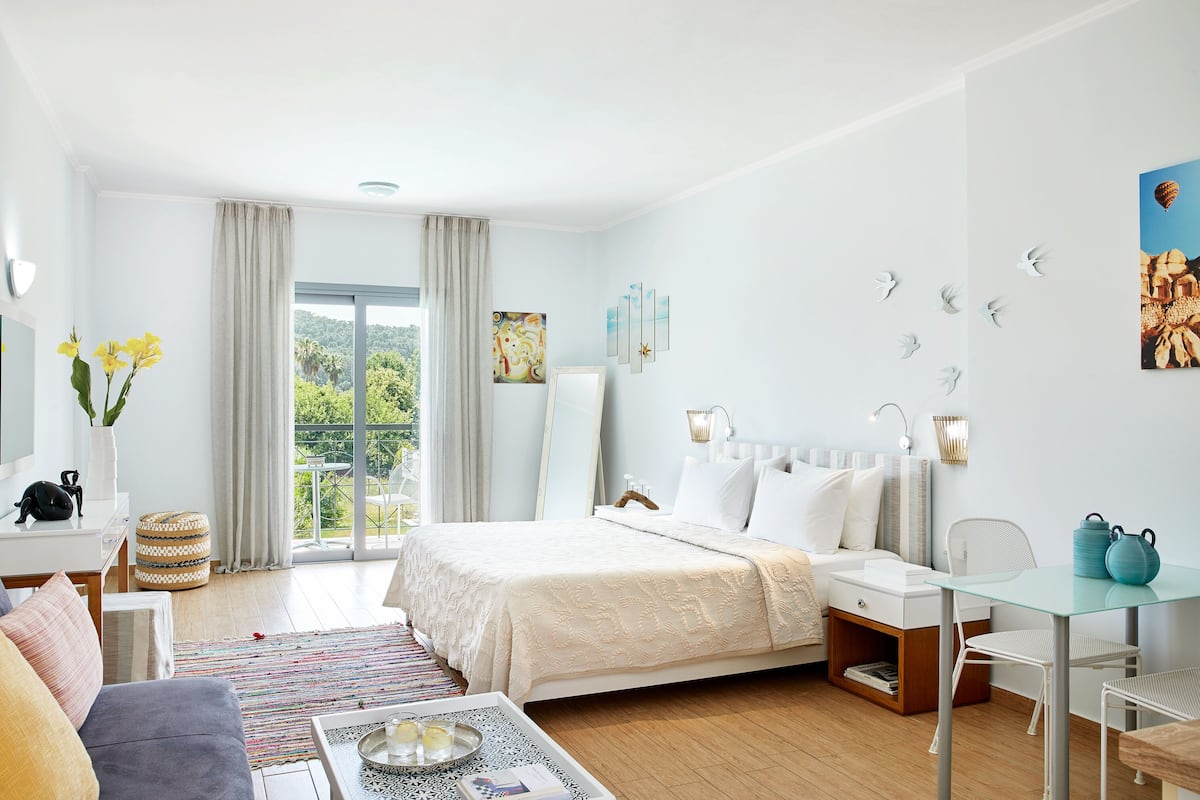A modern hotel room with a large bed, a sitting area with a sofa and a coffee table, a small desk, and a balcony with curtains.