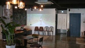 A modern dining area with a long table, several chairs, and hanging pendant lights. There is a projection screen displaying an image on the wall.