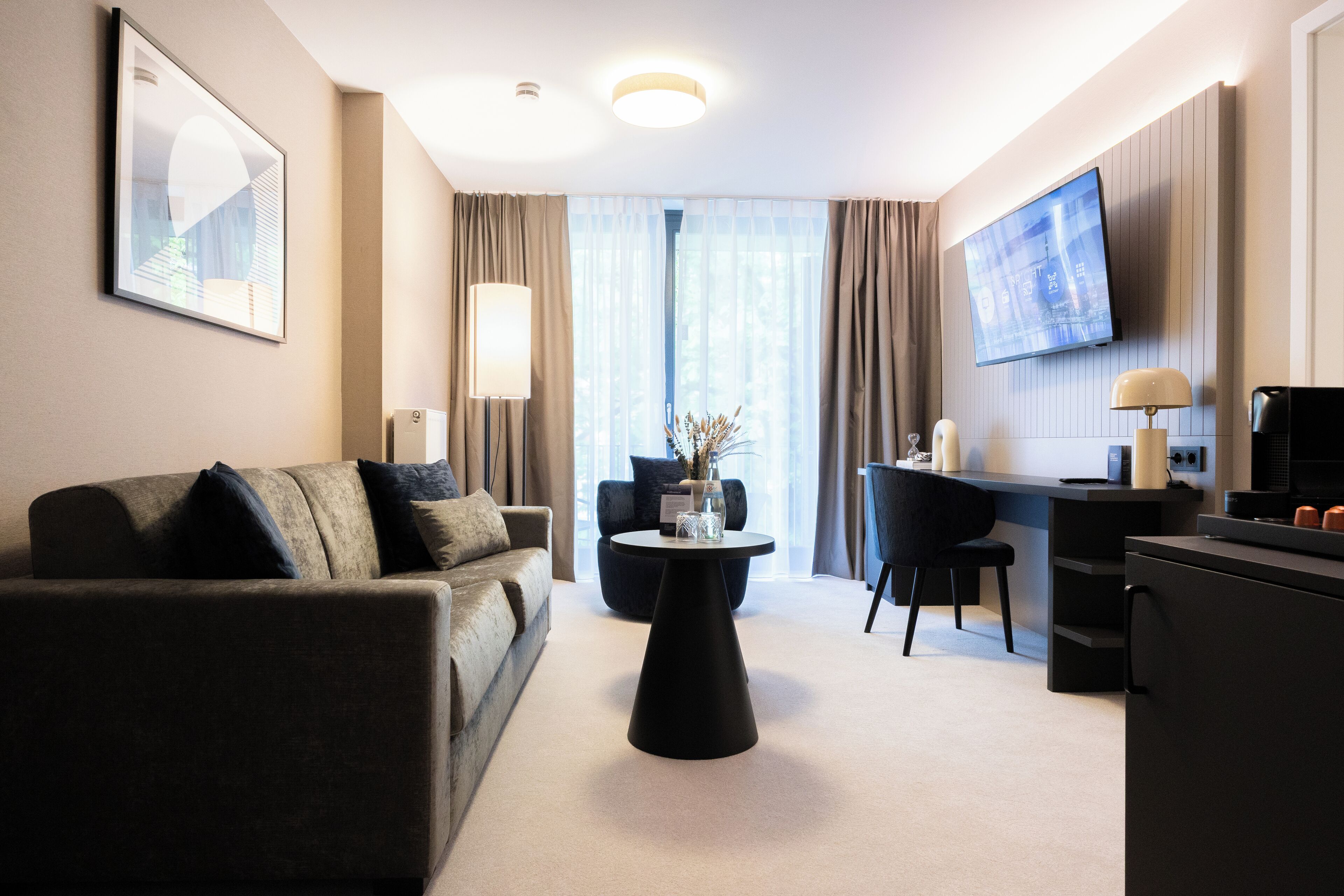 A modern hotel room with a sofa, a round coffee table, a desk, and a flat-screen TV mounted on the wall.