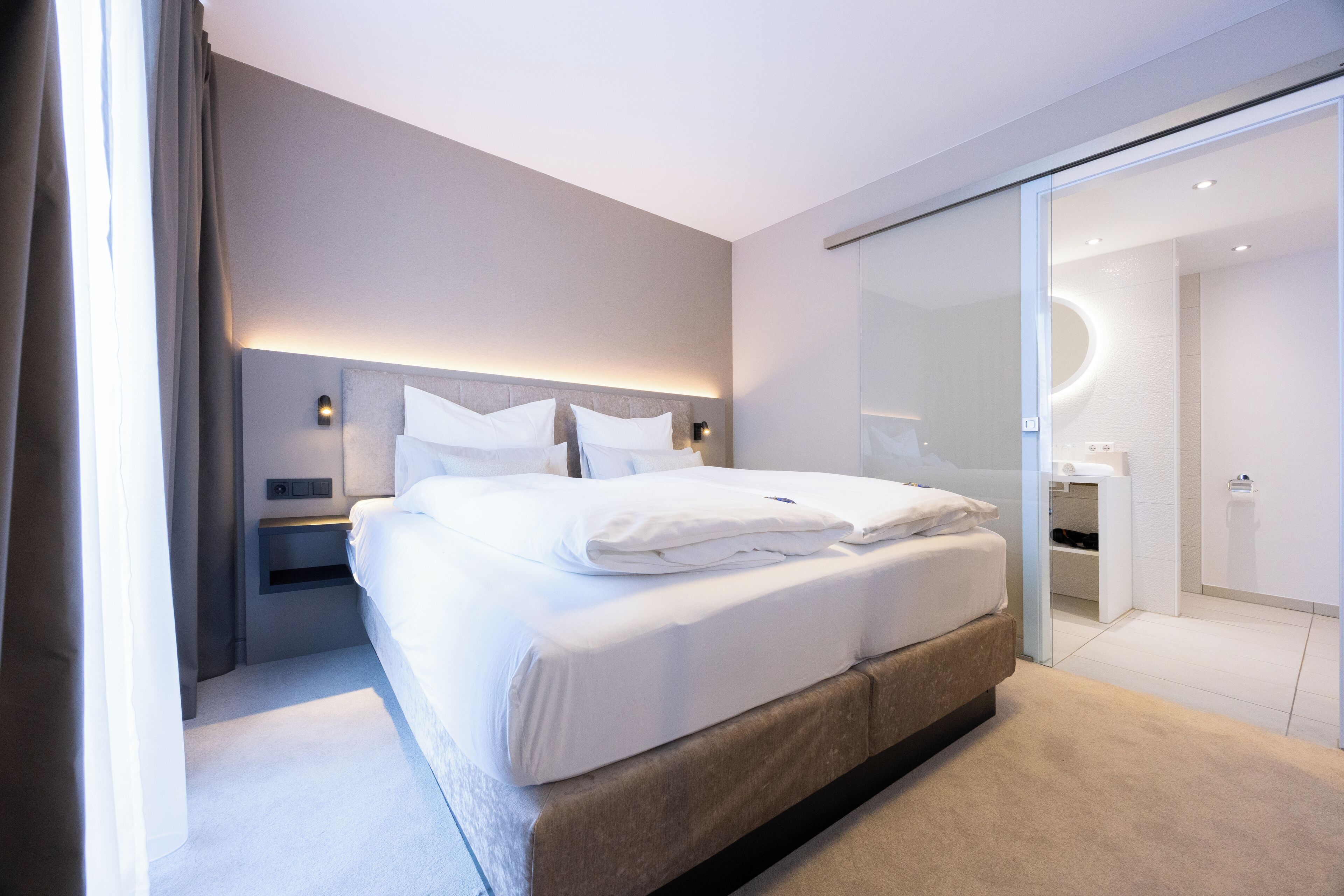 Ein modernes Hotelzimmer mit einem großen Bett, einem Nachttisch, einer Ablagefläche und einer Glasabtrennung zu einem Badezimmer.