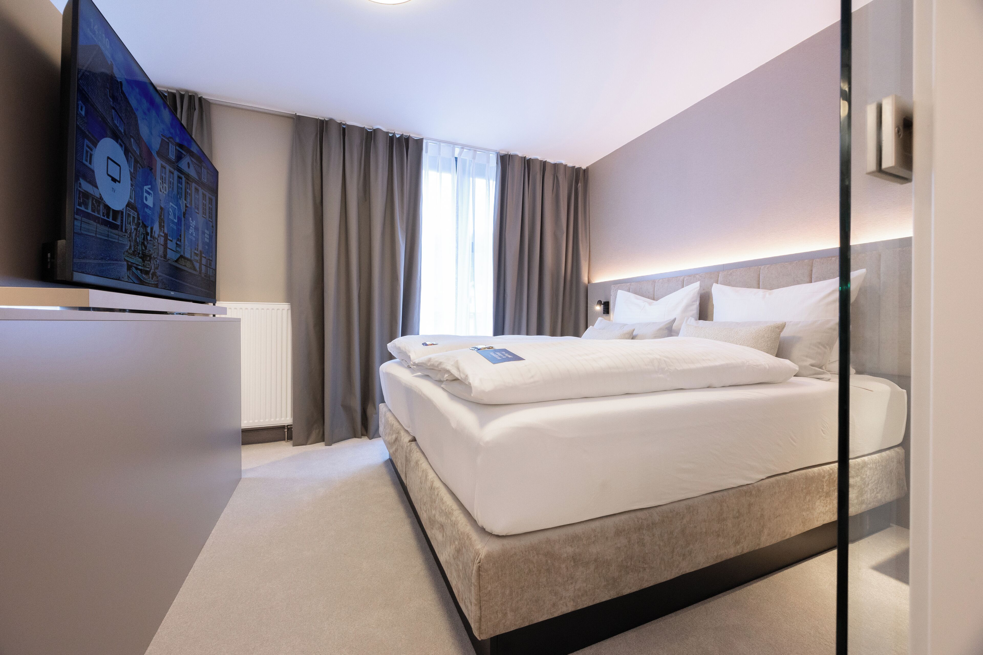 Habitación de hotel moderna con una cama grande, una televisión de pantalla plana y vistas de la ciudad a través de la ventana.