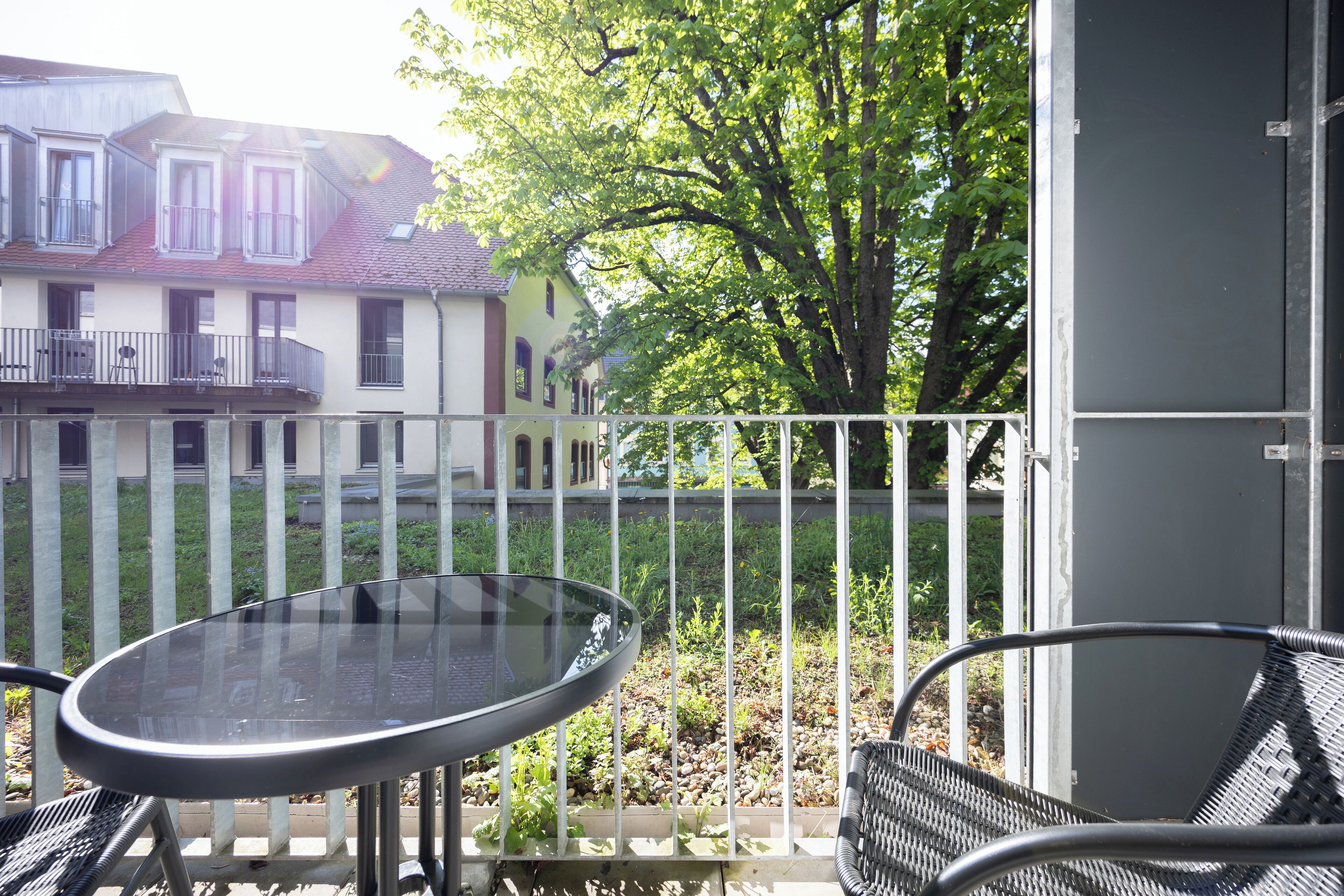 A balcony with a glass table and metal chairs, overlooking a garden and a building with a red roof.