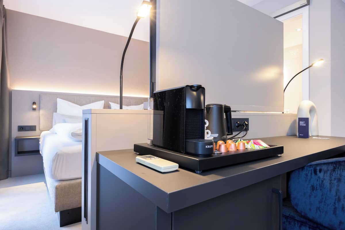 A modern hotel room with a bed, bedside table, and a desk with a coffee maker, kettle, and cups.