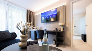 A modern hotel room with a flat-screen TV mounted on the wall, a sofa, a coffee table with a bottle and glasses, and a desk with a lamp. - Digital Hotel Kronjuwel by BRIGHT Waldkirch - Self Check-in (Waldkirch)
