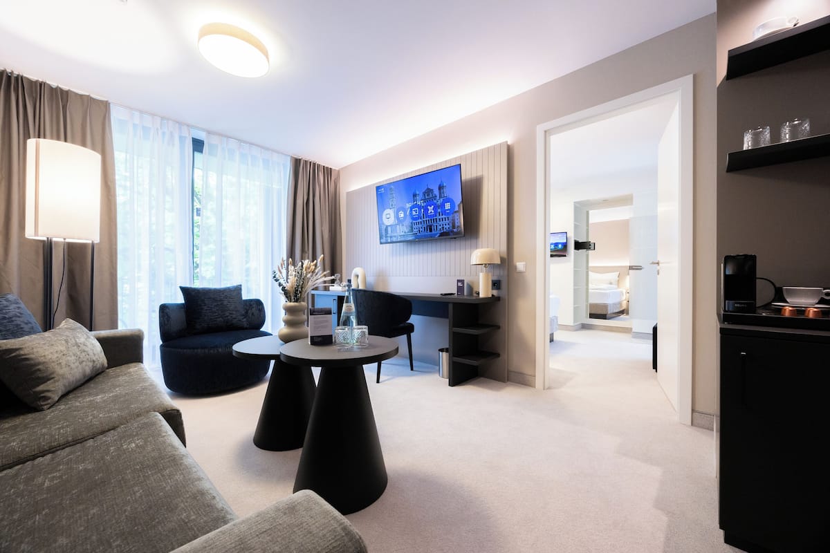 A modern hotel room with a sofa, a round coffee table, a flat-screen TV mounted on the wall, a desk with a chair, and a kitchen area.