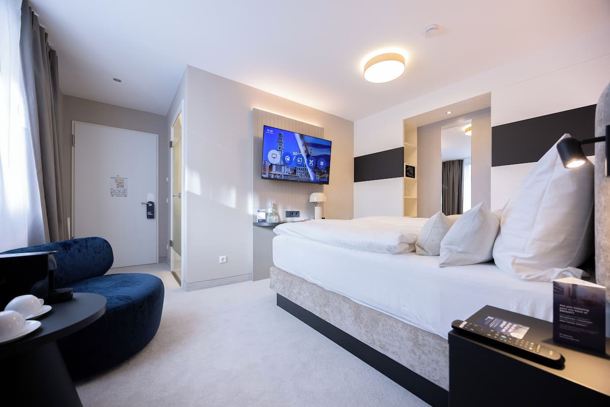 A modern hotel room with a large bed, a flat-screen TV mounted on the wall, a bedside table with a remote control, and a blue accent chair.