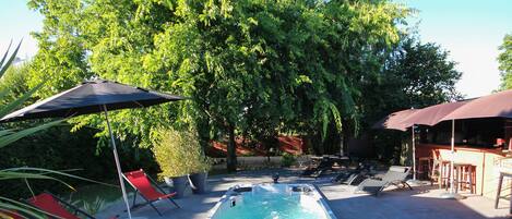 A rectangular hot tub with a pool, surrounded by red lounge chairs and umbrellas, set in a garden with trees and a barbecue area.