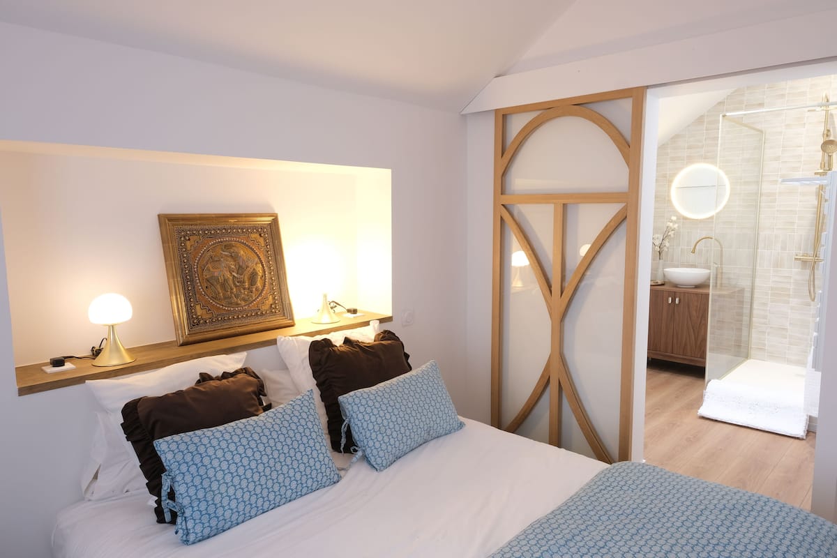 Un dormitorio con cama, lámparas de noche, una obra de arte en la pared y un baño con ducha y lavamanos.