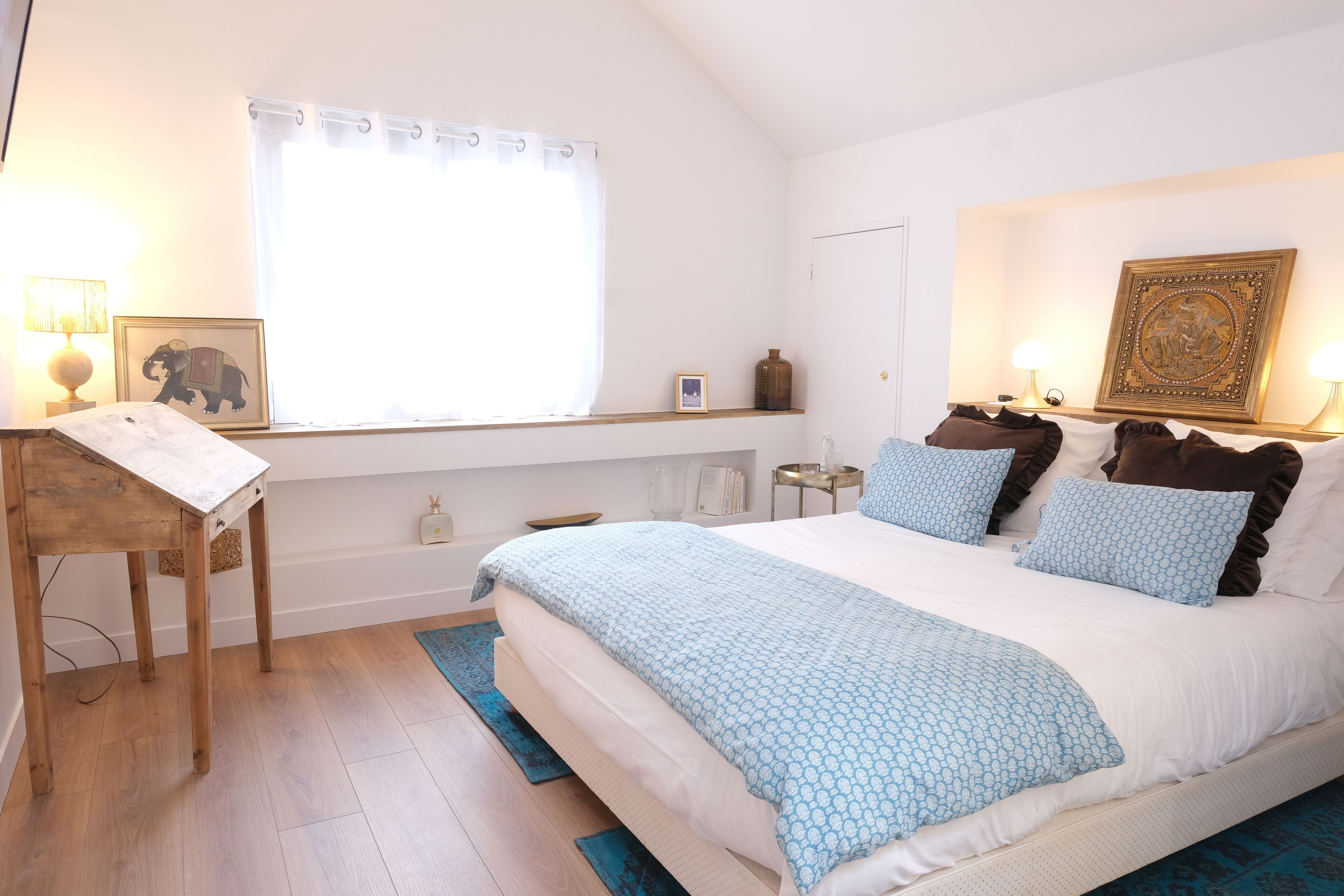 Un dormitorio con una cama grande, una mesita de noche de madera, un ventanal con cortina y un estante en la pared con adornos.