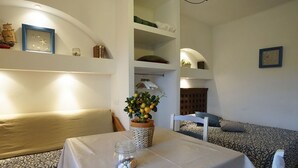 Una habitación acogedora con una mesa de comedor, un banco y un sofá. Hay estantes con diversos objetos y un reloj en la pared.
