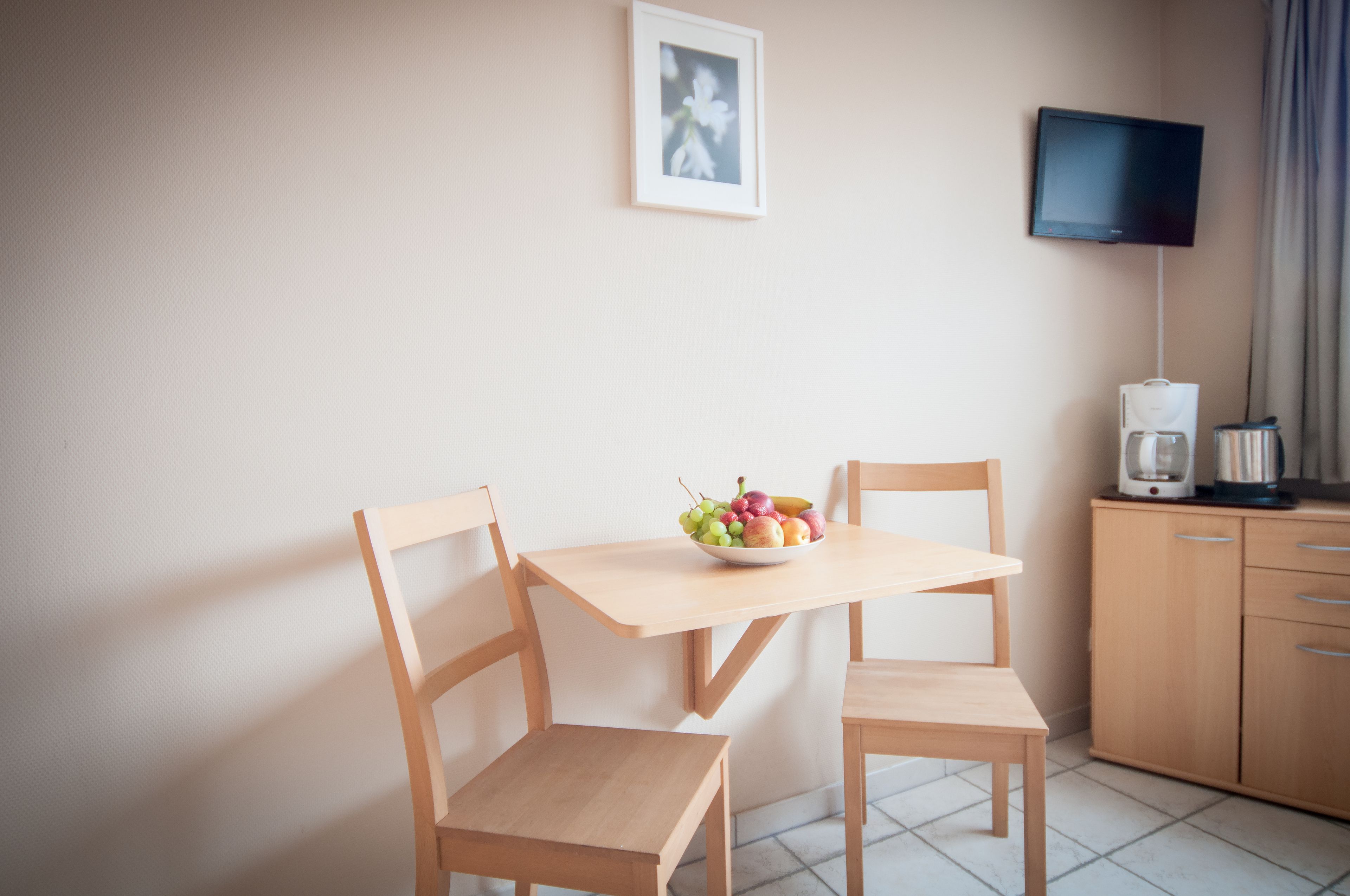 A small, clean room with a wooden table, two chairs, a framed picture on the wall, a television, and a coffee maker on a cabinet.