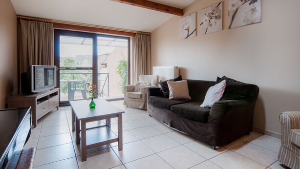 A living room with a sofa, a TV, a table, and a chair. There is a window with curtains and a view of the outside. - Holiday Home 't Regulierenhuisje (Tongeren)