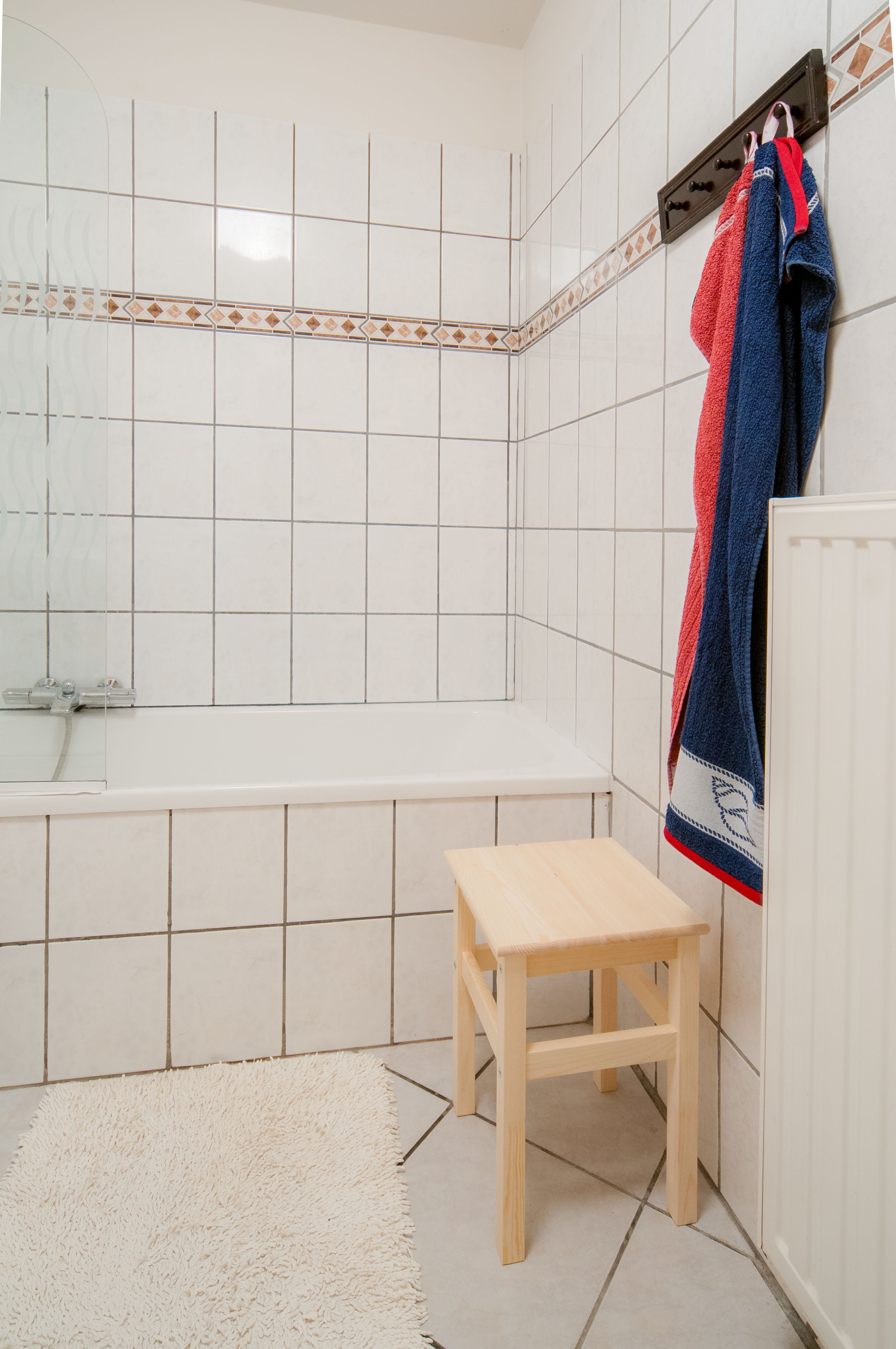 Une salle de bain avec des murs recouverts de carreaux blancs, un tabouret en bois et un tapis blanc.