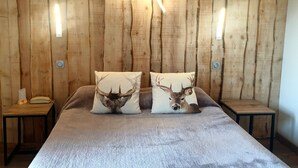 Una habitación con pared de madera, cama con dos almohadas decoradas con cuernos de ciervo, mesita de noche con teléfono, y lámpara moderna.
