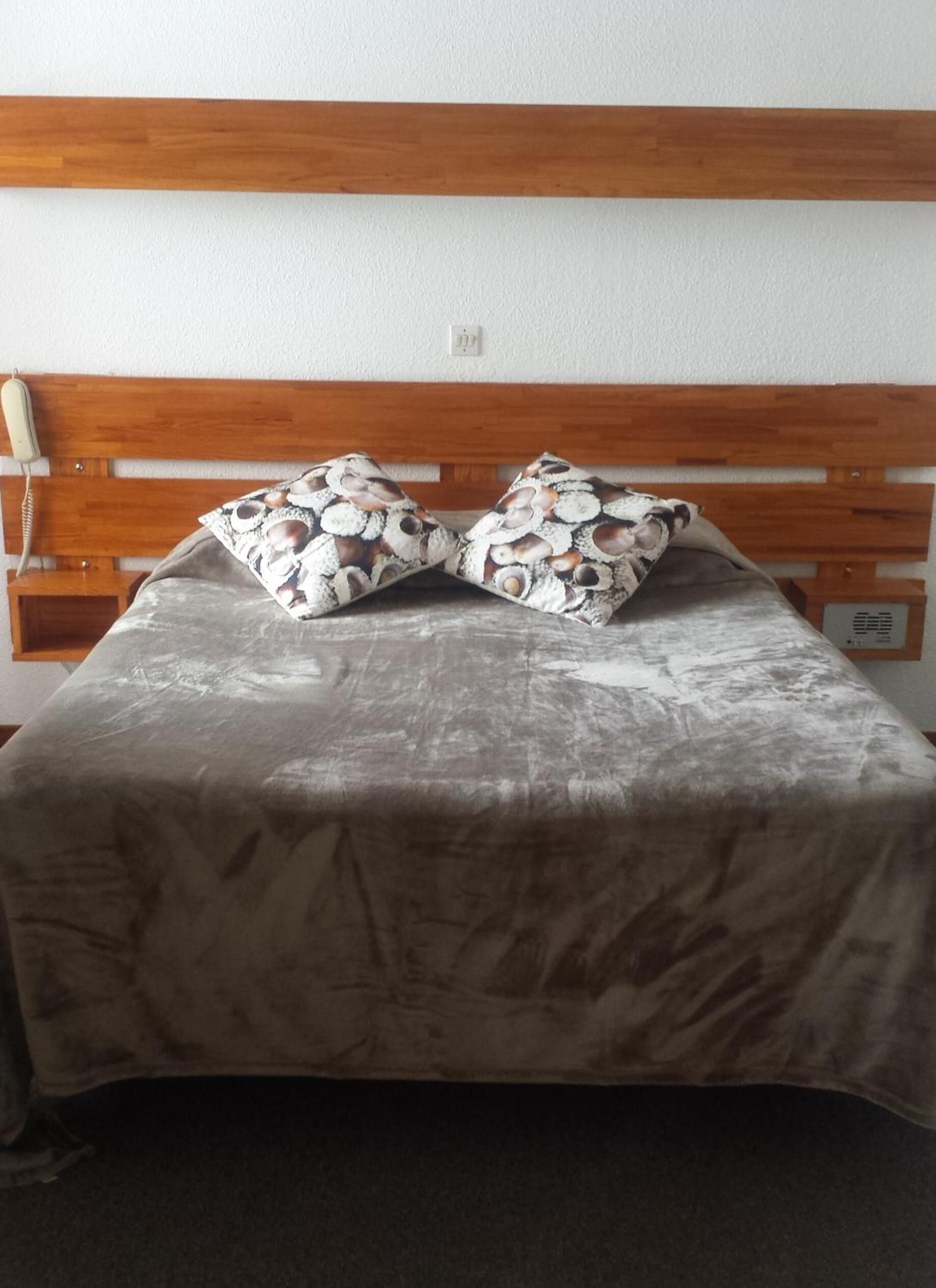 Un lit avec une tête de lit en bois, deux oreillers à motifs de coquillages et une couvre-lit grise.