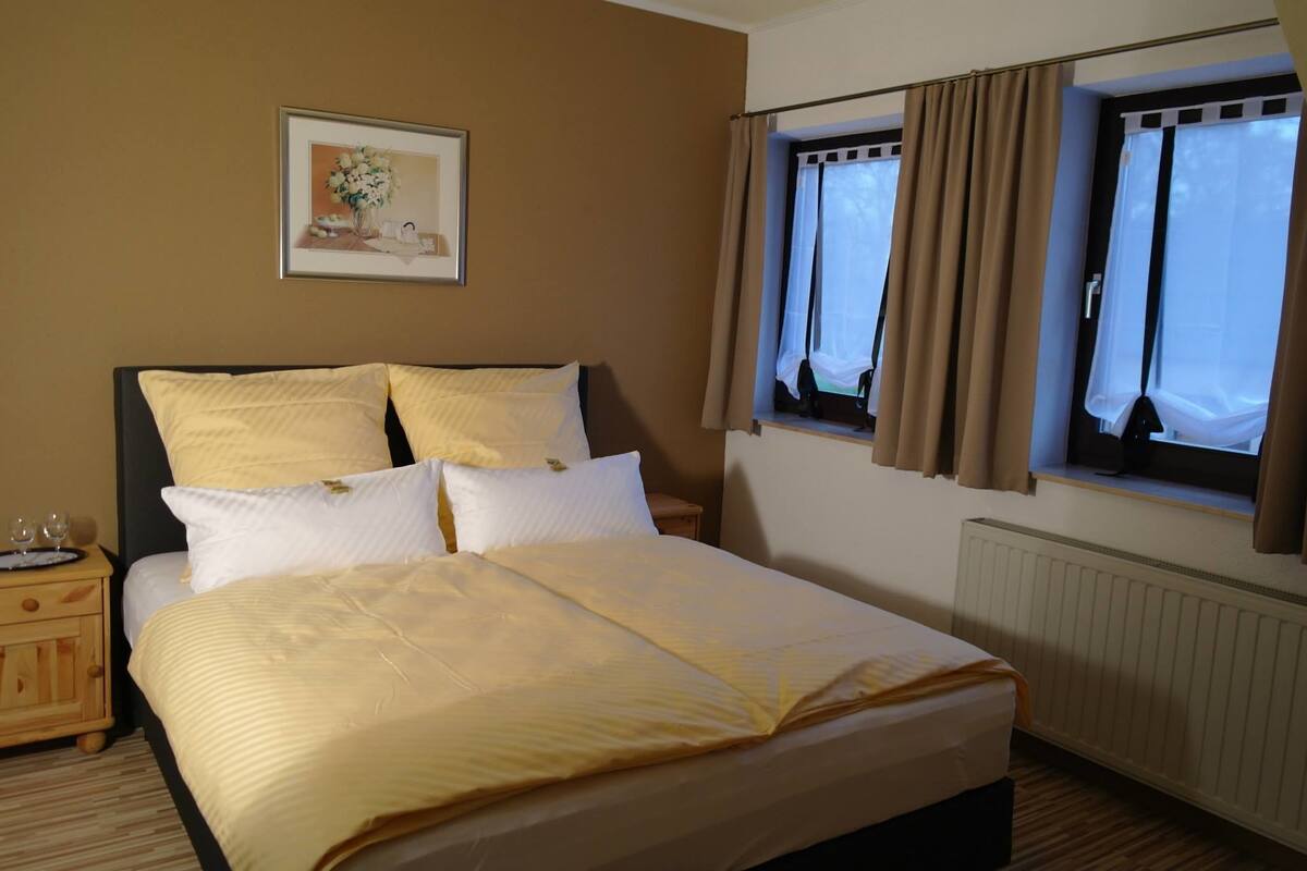 A neatly made bed with a yellow bedspread and pillows, a bedside table with a glass, and a framed picture on the wall.