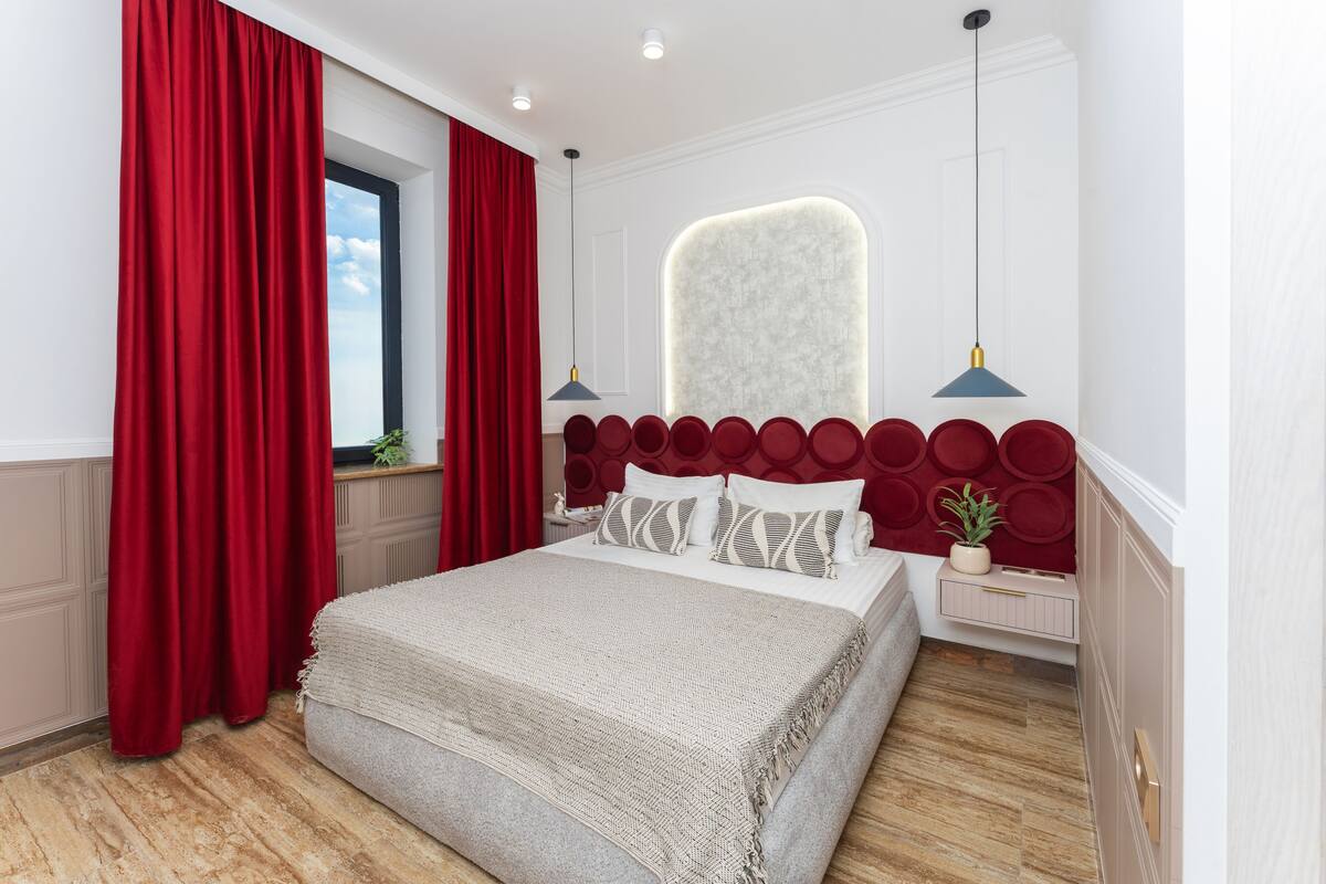 A bedroom with a bed, red curtains, a window, a bedside table with a plant, and a wall-mounted light fixture.