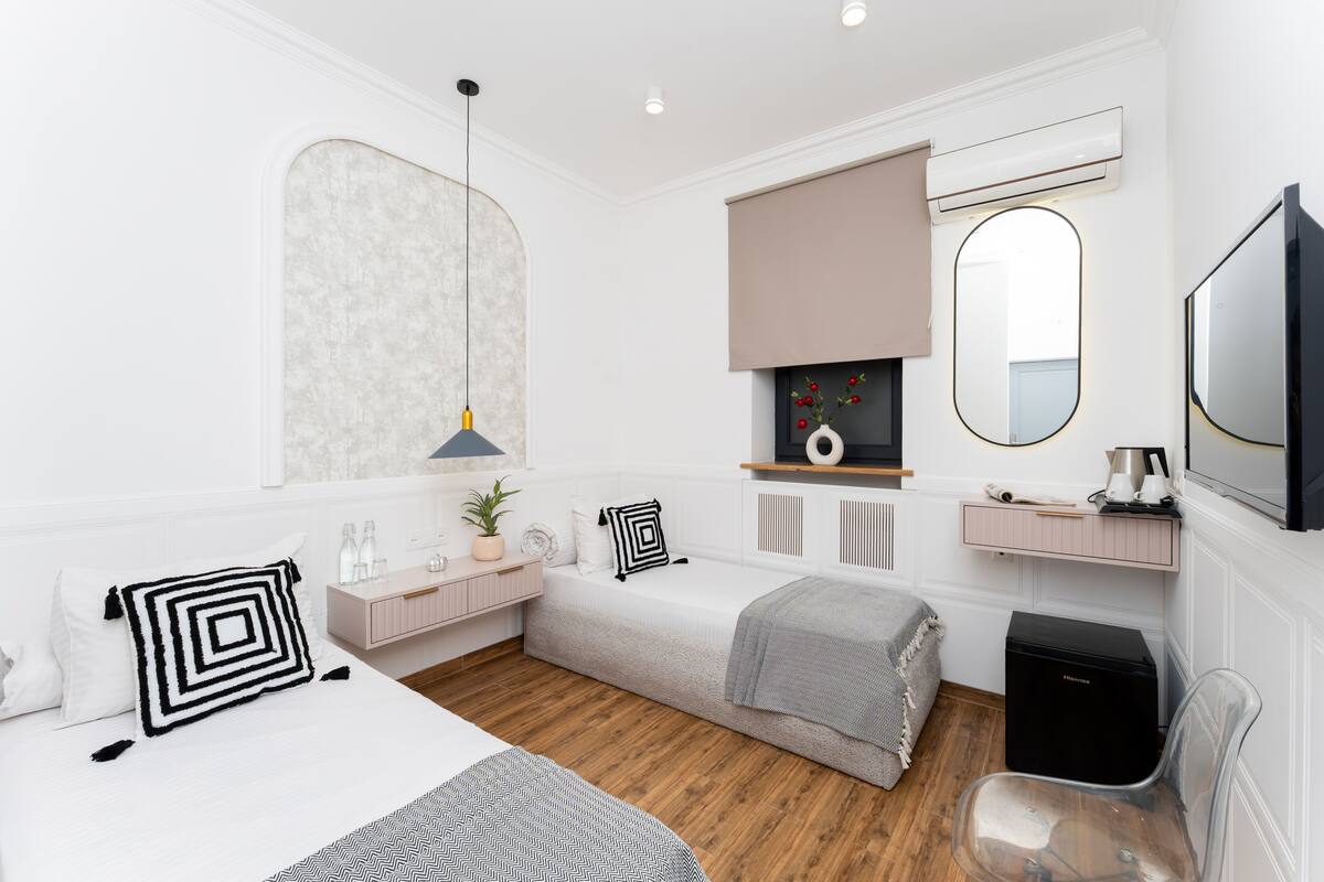 A modern bedroom with a bed, a desk, a chair, a small sofa, a mirror, and an air conditioning unit.