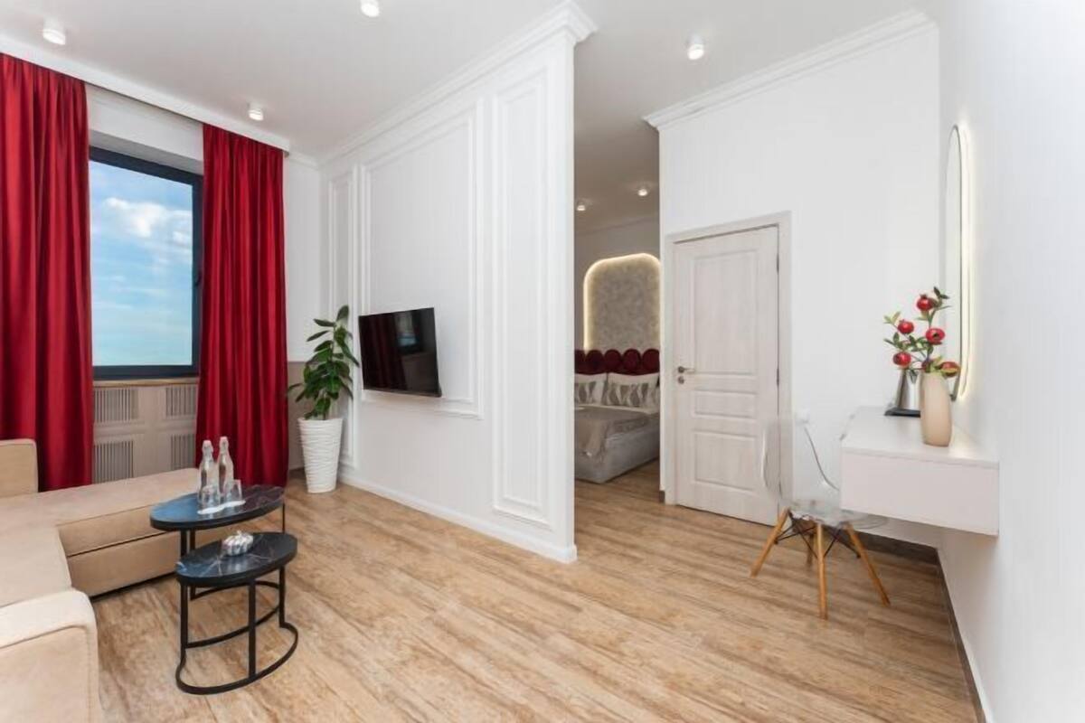 A modern living room with a sofa, a coffee table, a TV, and a large window with red curtains.