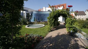 A garden with a paved walkway, a fountain, and a modern building with a glass facade.