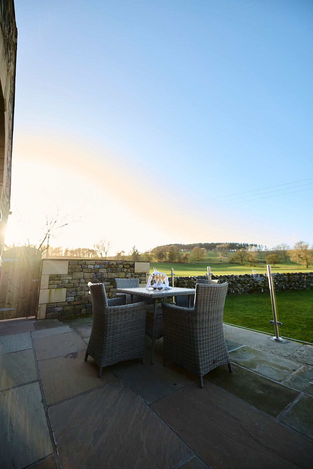An outdoor patio with wicker furniture and a table set for two, overlooking a grassy landscape.
