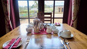 A dining table set with white dishes, silverware, and a silver teapot, with a view of a garden through a window. - GreenGrove Country House B&B (Skipton)