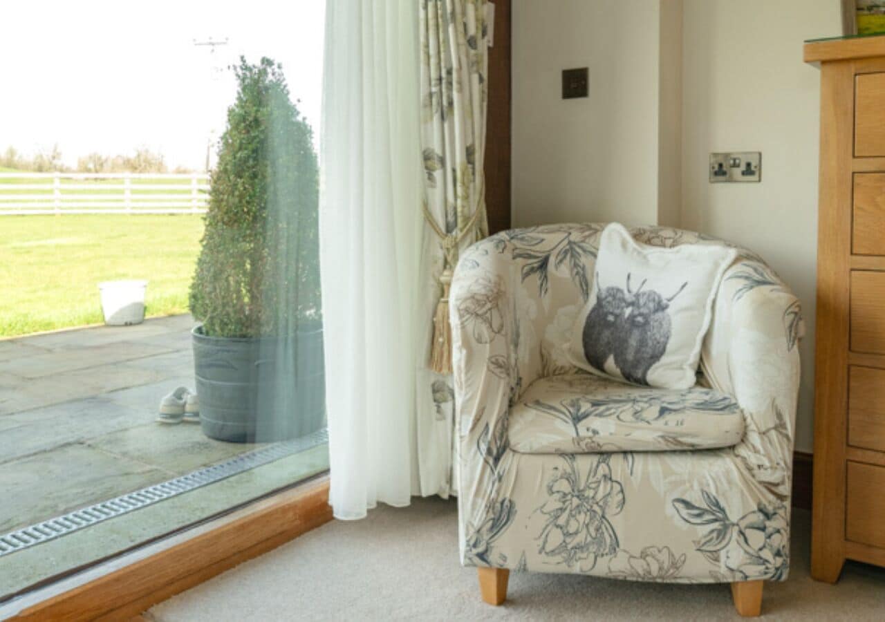 A room with a floral-patterned armchair, a potted plant, a wooden cabinet, and a view of a grassy field through a window.