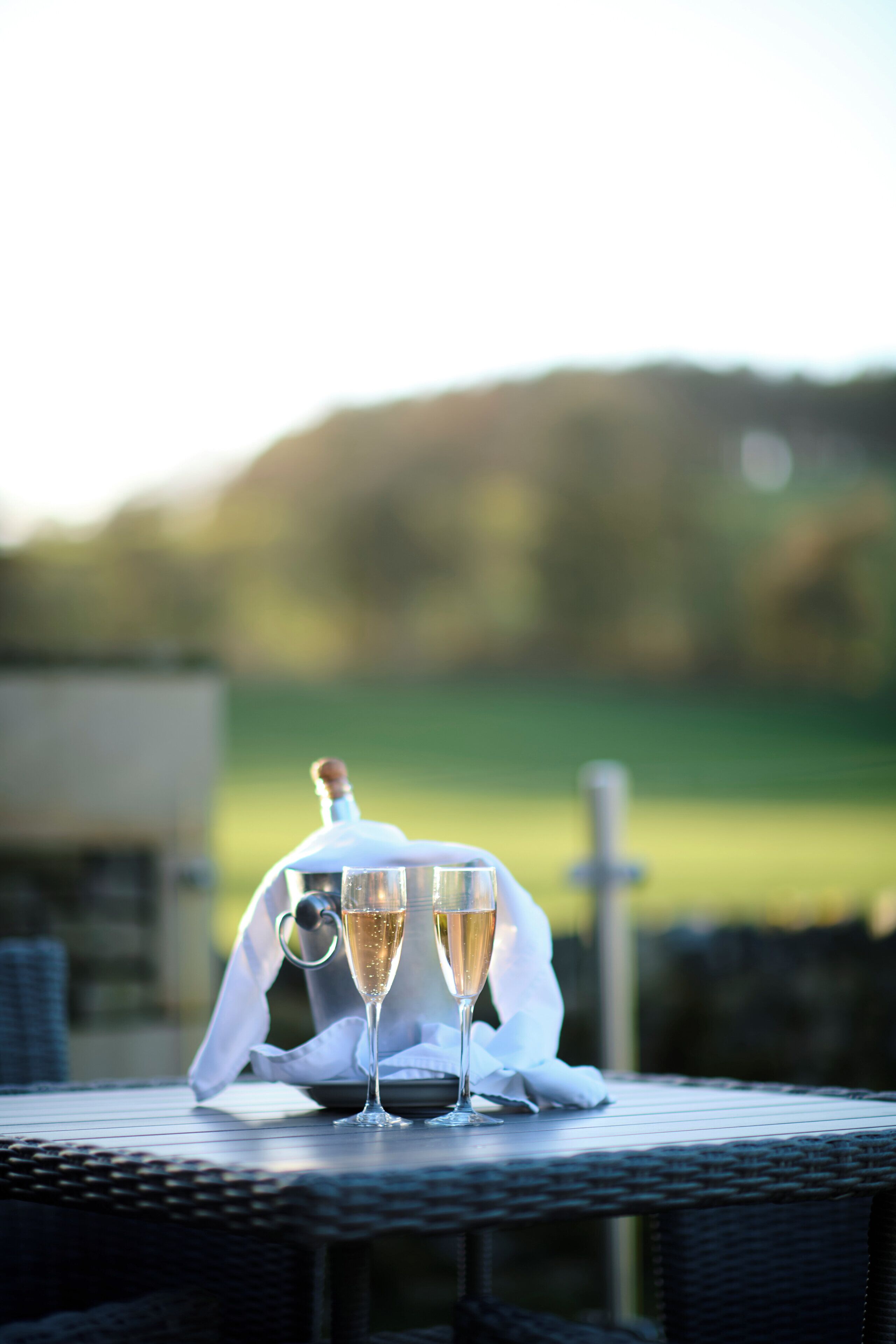 A table with two champagne glasses and a bottle, set on a wicker table outdoors with a blurred green landscape in the background.