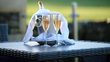 A table with two champagne glasses and a bottle, set on a wicker table outdoors with a blurred green landscape in the background.