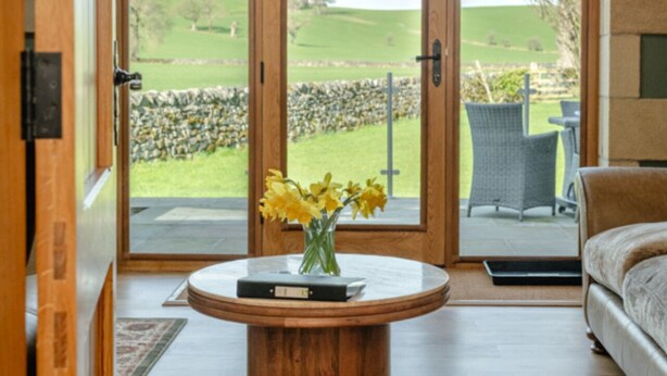 A room with a wooden door, a round table with a book and flowers, a sofa, and a view of a green landscape through glass doors. - GreenGrove Country House B&B (Skipton)