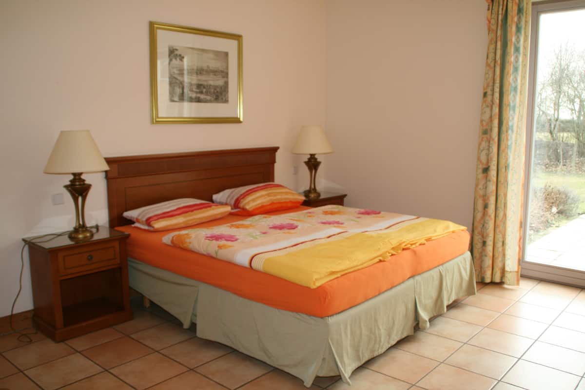 Un dormitorio con una cama, dos mesitas de noche, una mesita auxiliar, un cuadro en la pared y una ventana con cortinas.