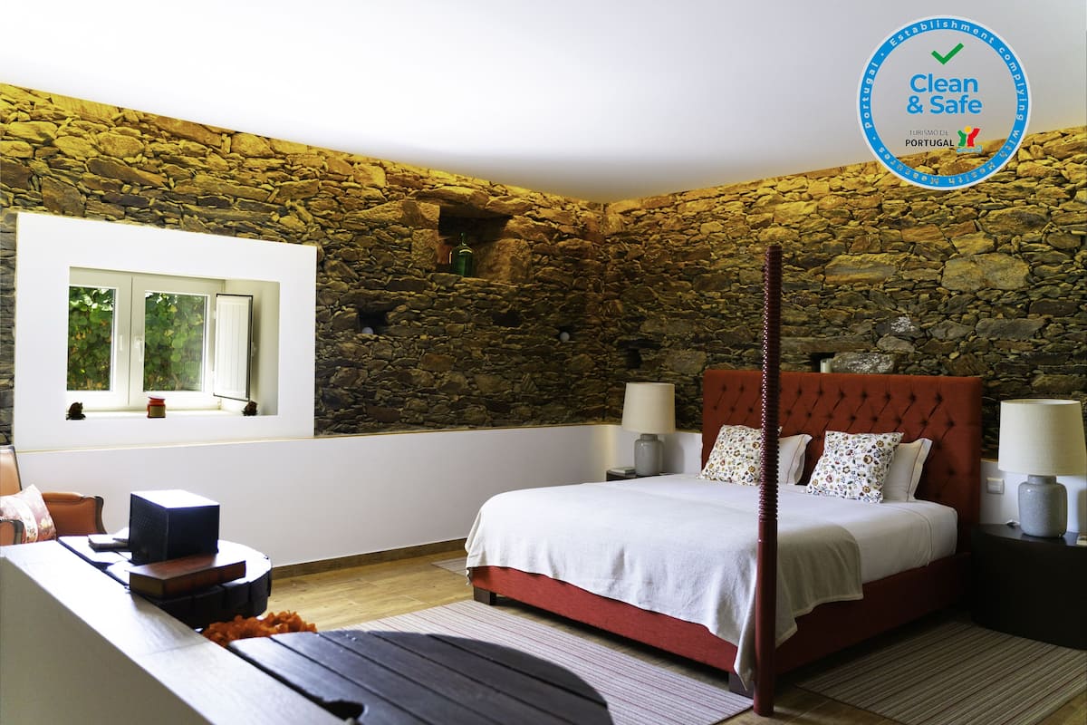 Une chambre avec un mur en pierre, un lit avec une tête de lit rouge, des lampes de chevet et une fenêtre donnant sur un paysage verdoyant.