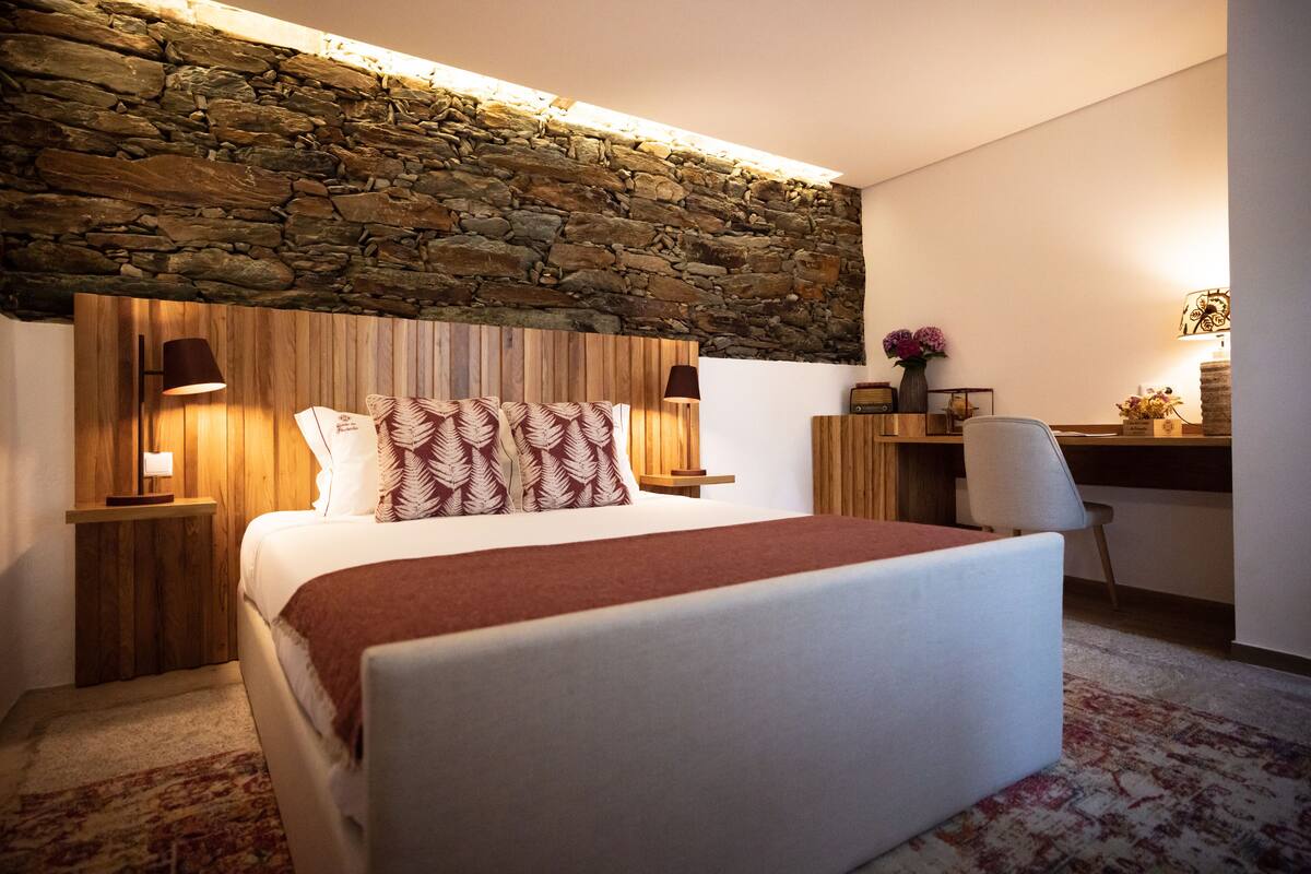Une chambre avec un mur en pierre, un lit recouvert d’une couverture rouge, deux lampes de chevet et un bureau avec une chaise.