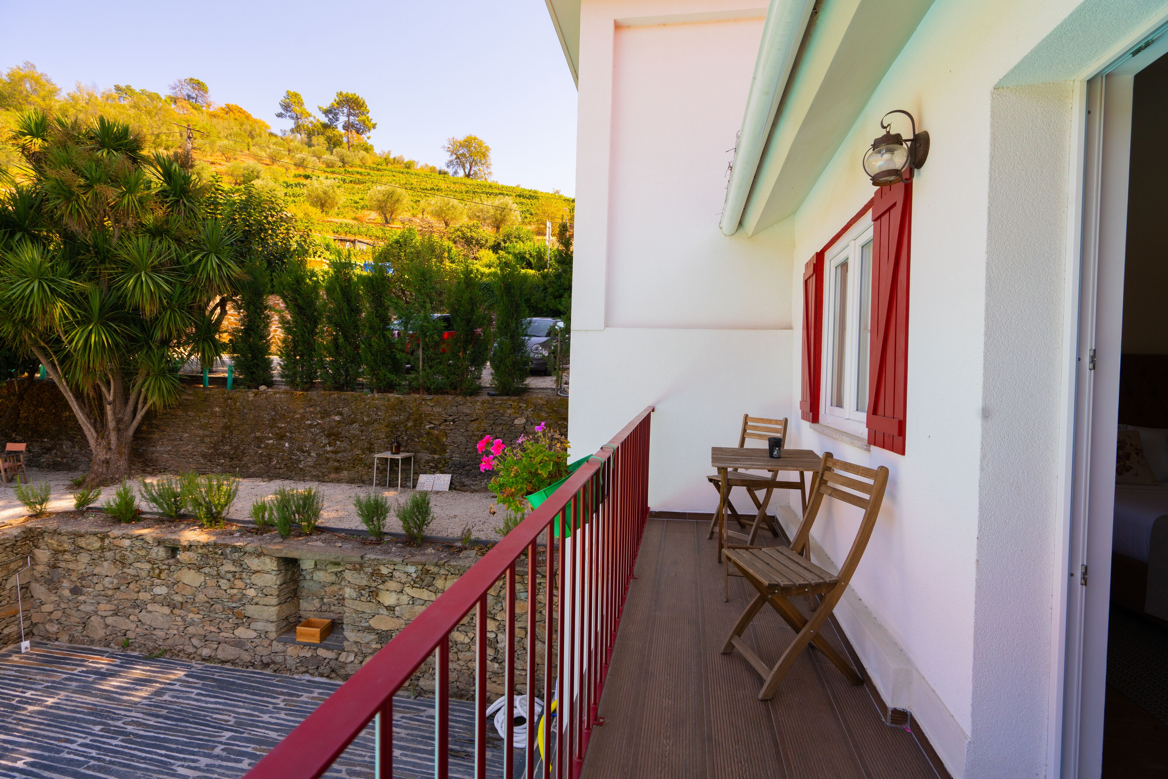 Un balcón con pared de piedra, una mesa y sillas de madera, y vistas a una colina con árboles y coches aparcados.