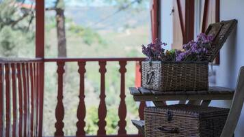 Un balcón con cestas de mimbre con flores moradas, una mesa de madera y vistas a zonas verdes y colinas.