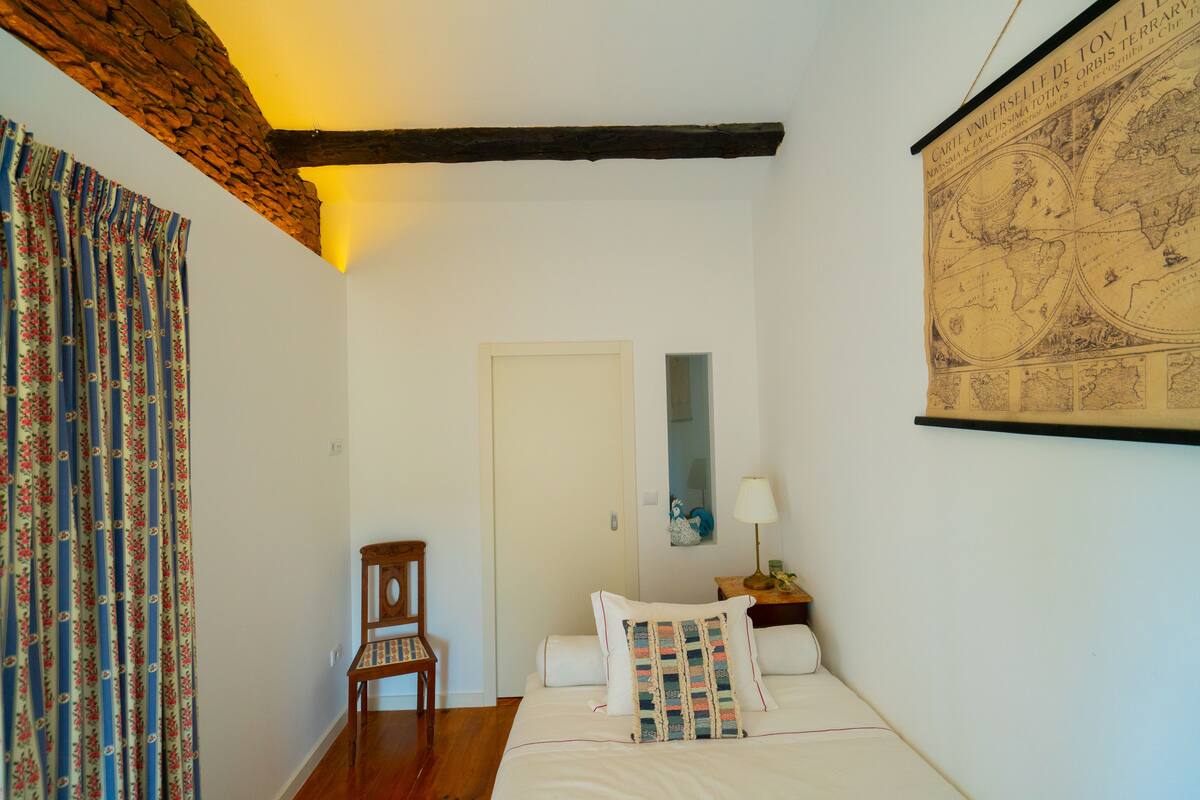 Une chambre à coucher avec un lit, une chaise en bois, une table de chevet avec une lampe, une carte accrochée au mur et une porte.