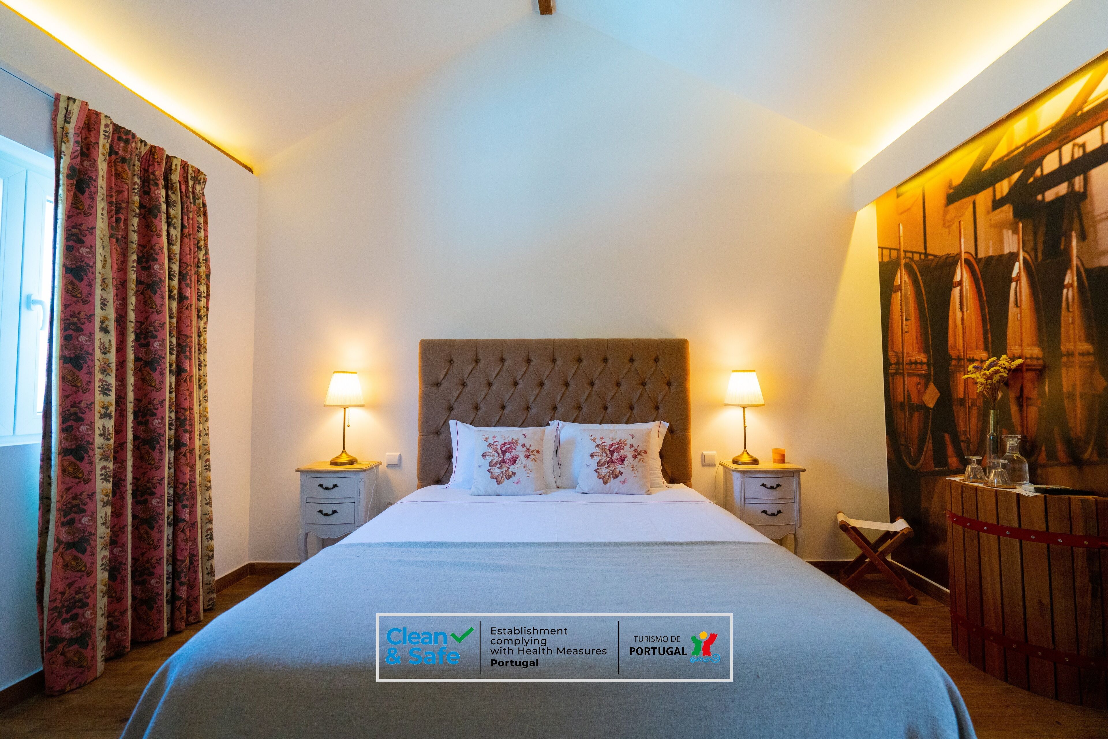 Habitación de hotel con una cama bien hecha, dos mesitas de noche con lámparas, cortinas florales y una pared con un barril de madera y un cuadro.