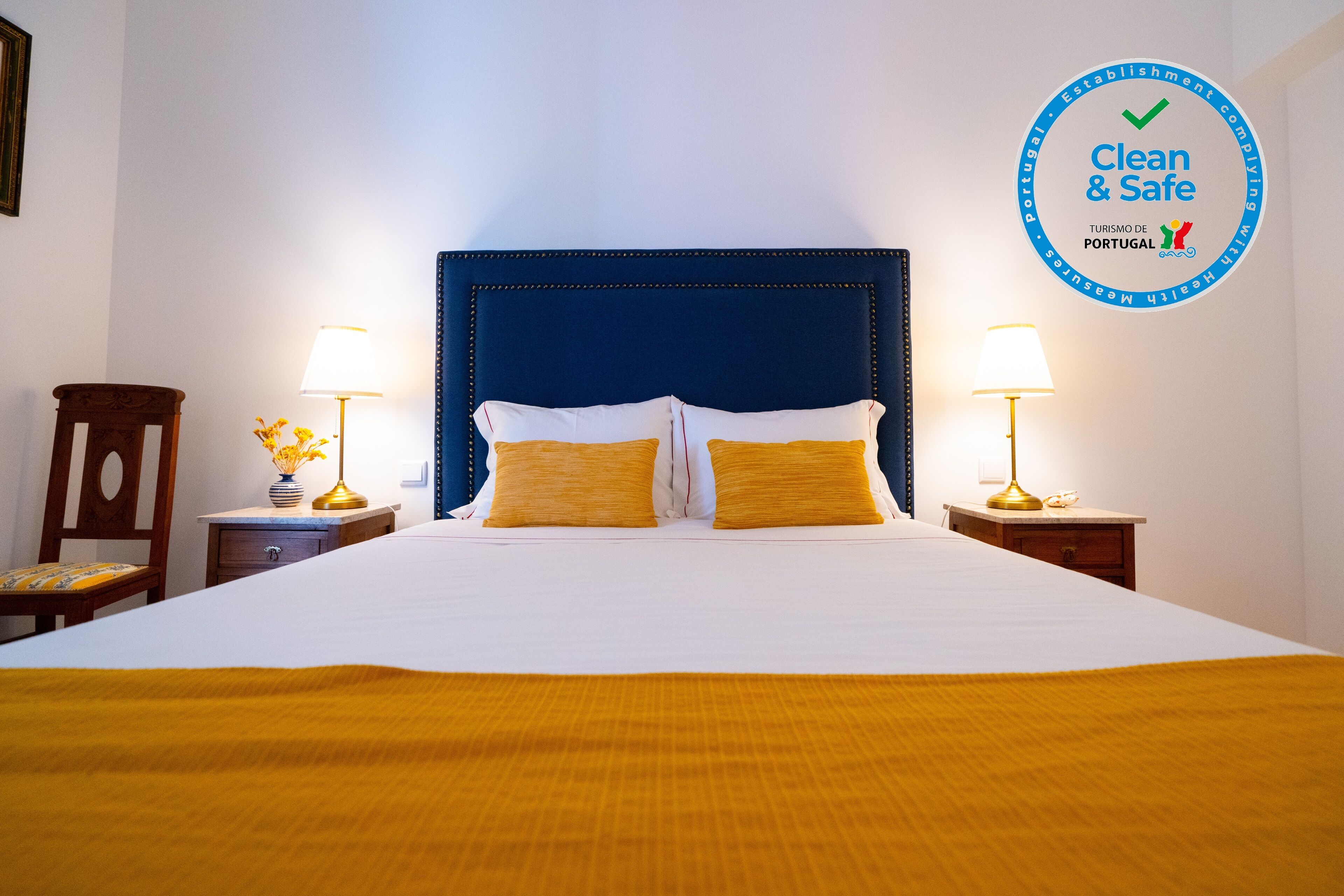 A hotel room with a large bed, two bedside tables with lamps, a wooden chair, and a wall sign indicating 'Clean & Safe' with a checkmark.