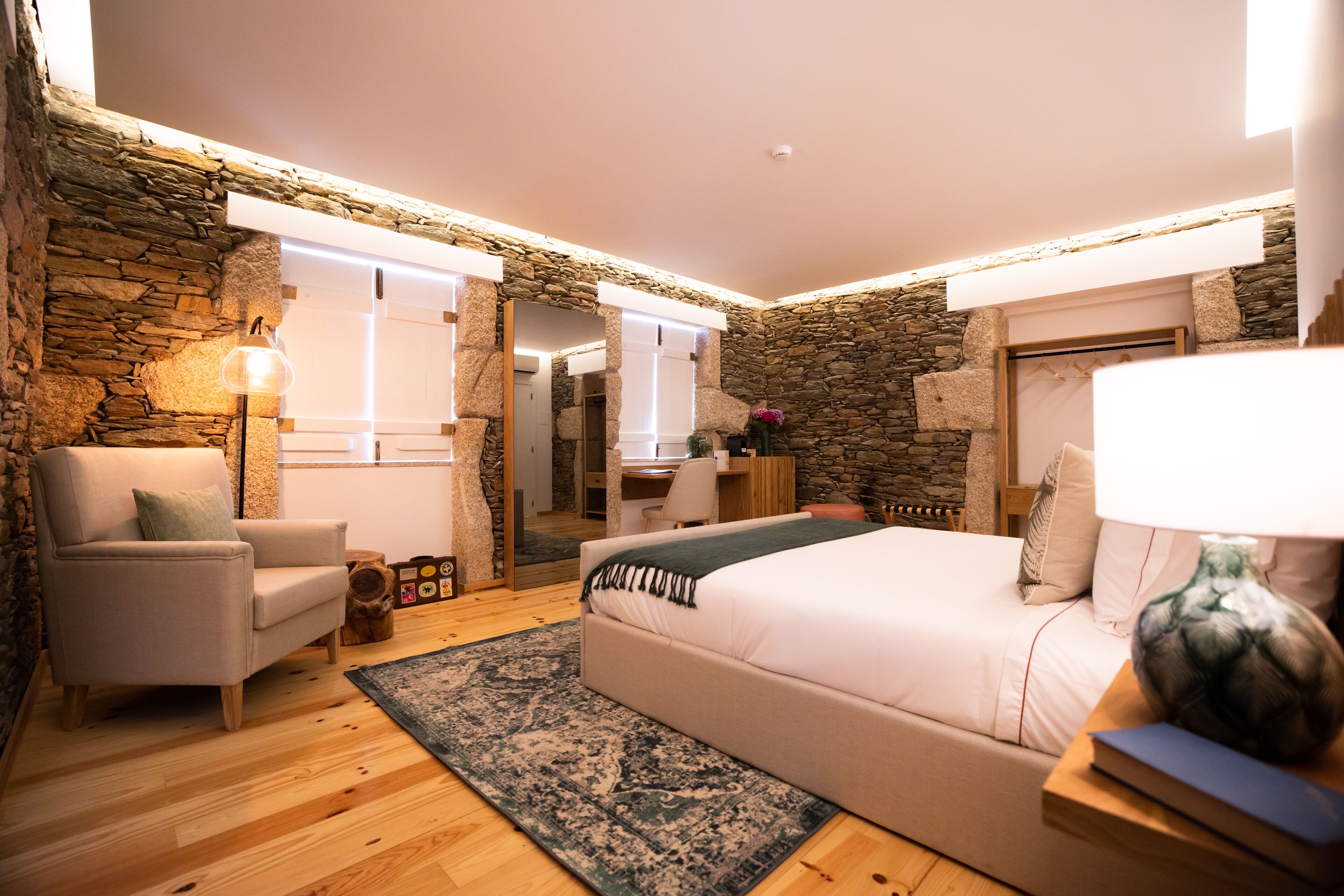 A bedroom with a stone wall, a bed, a chair, a desk, and a lamp.