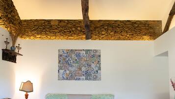 Ein gemütliches Zimmer mit Steinwand, Holzbalkendecke, einem gemusterten Teppich, einer Couch und einem Tisch mit Lampe.