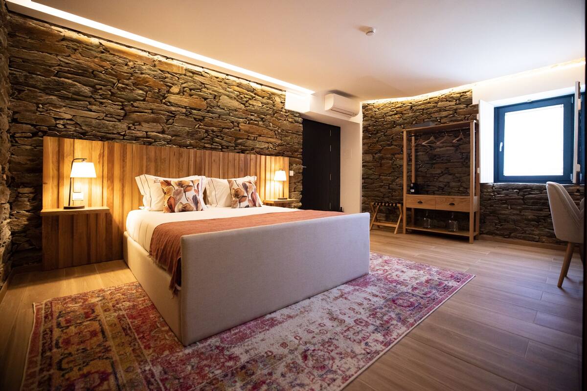 Une chambre avec un mur en pierre, un lit avec une tête de lit, des lampes de chevet, une armoire en bois et une fenêtre laissant entrer la lumière du jour.