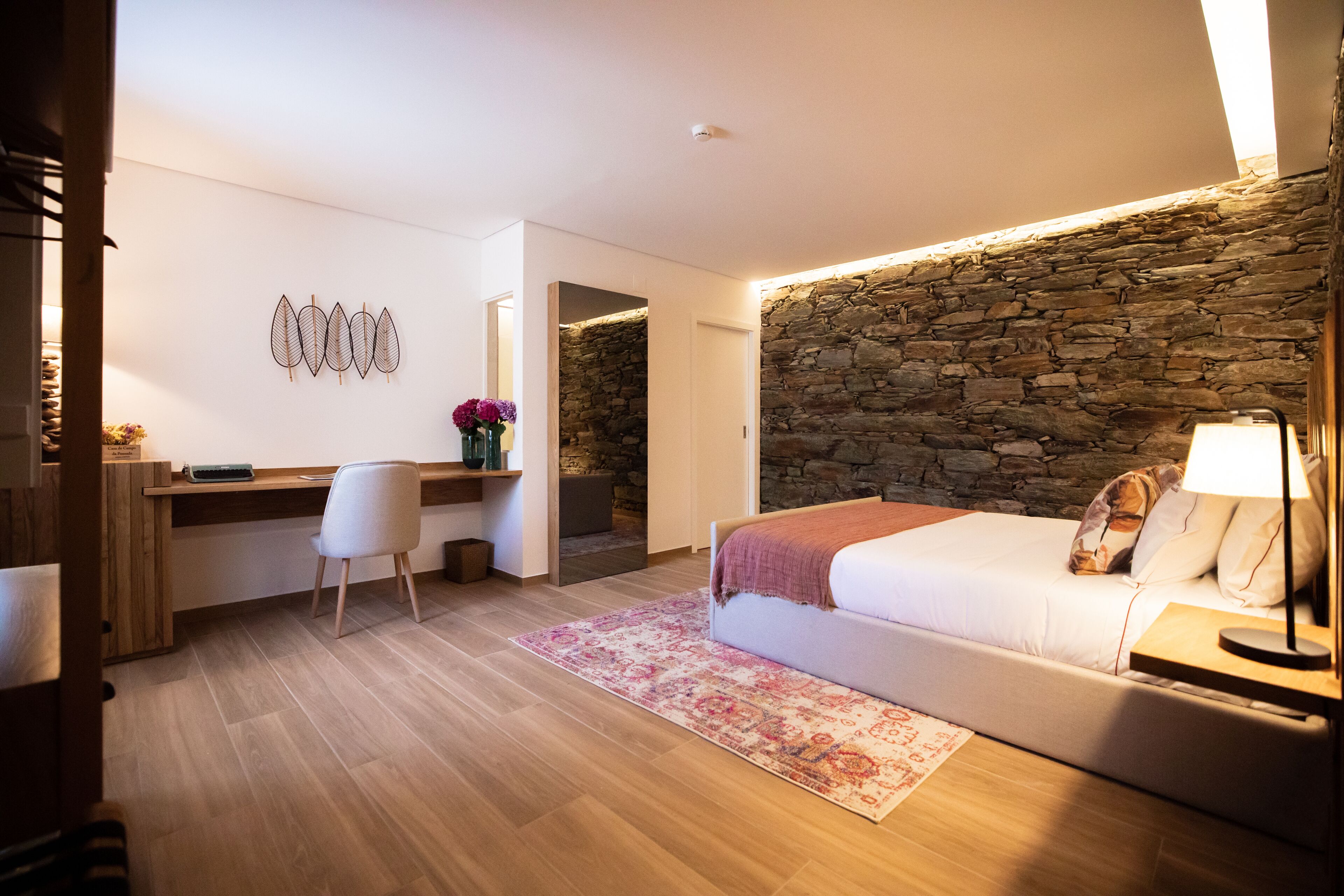 Une chambre moderne avec un mur d’accent en pierre, un lit avec une lampe, un bureau avec une chaise et un petit coin salon.