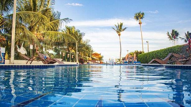 A swimming pool with clear blue water, surrounded by palm trees and lounge chairs. - Pousada Polymar (Maragogi)