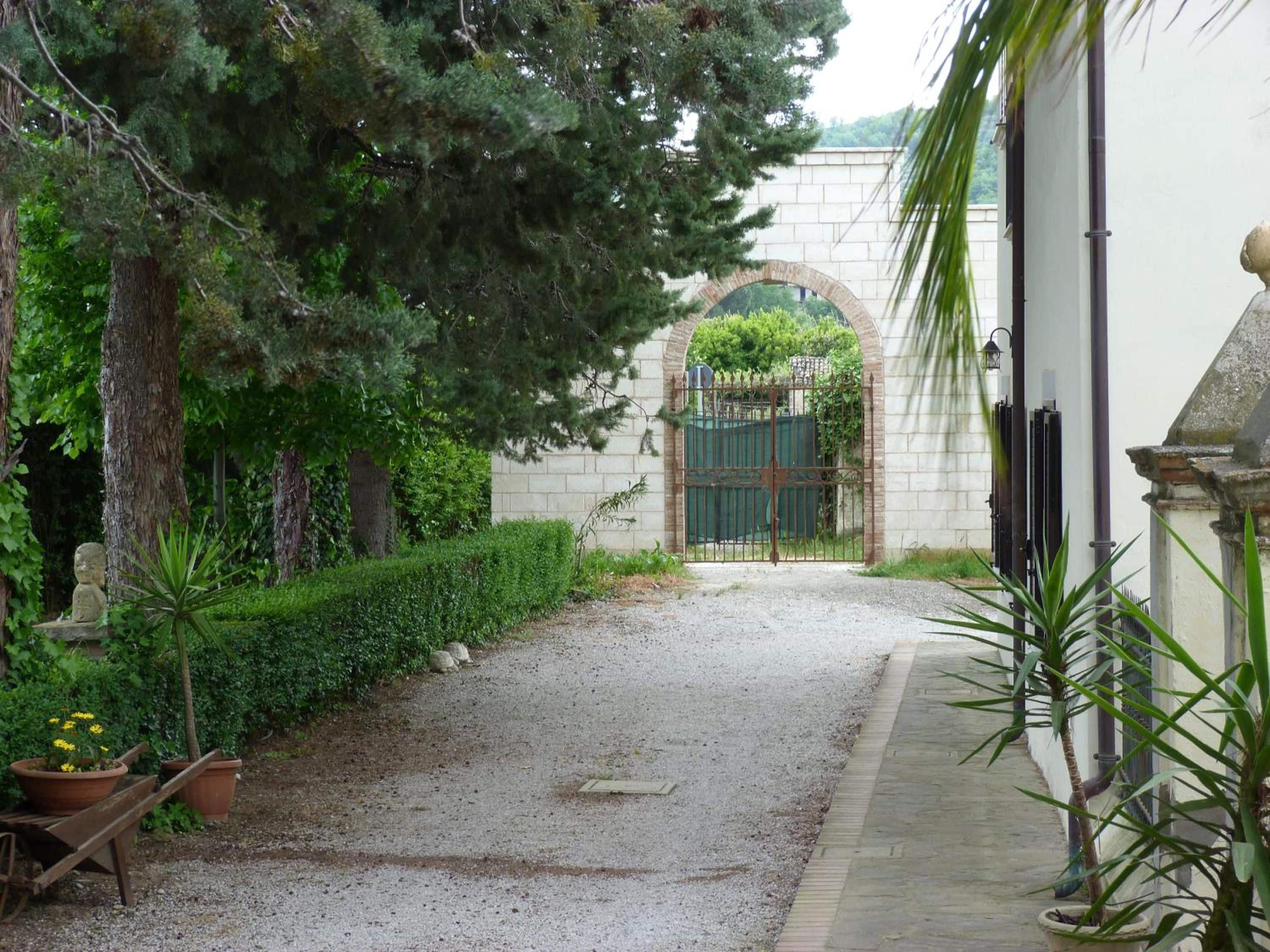 Un chemin de gravier mène à une entrée fermée par un portail surmonté d’une arche en pierre, le tout étant entouré d’une végétation luxuriante et de plantes en pot.